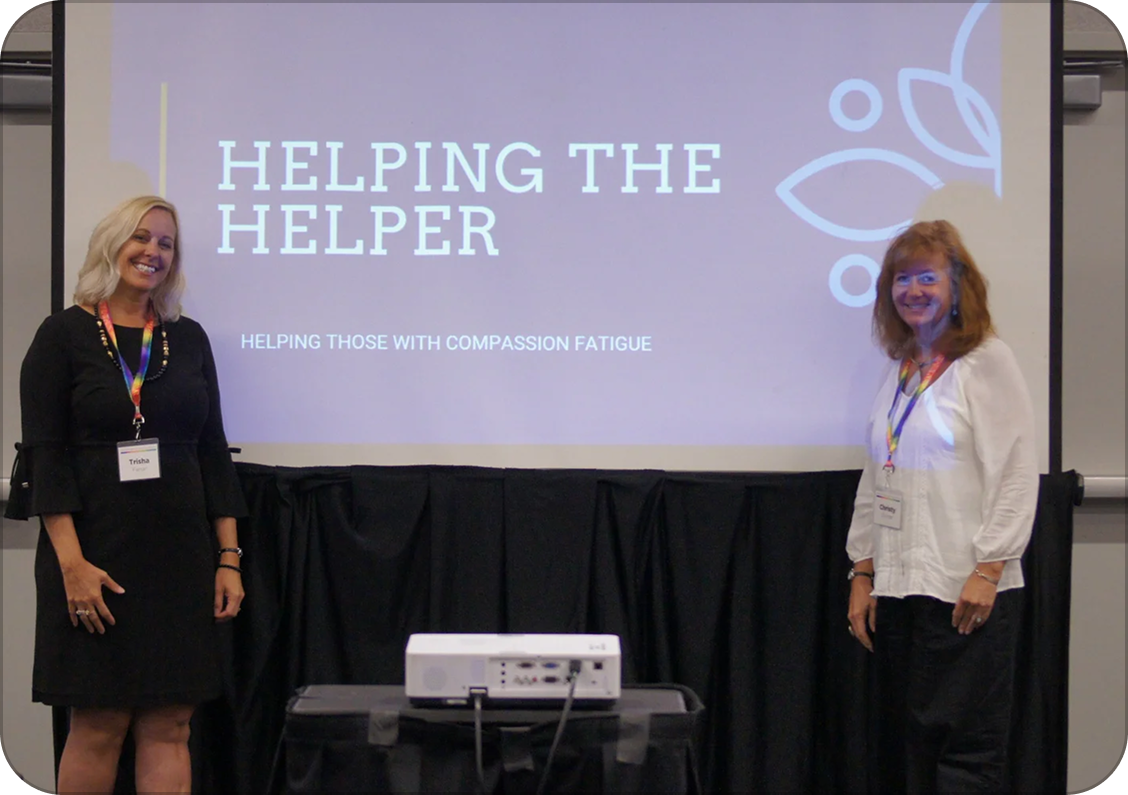 Trisha and Christy standing on either side of a projection screen with the text "Helping the Helper" and "Helping those with compassion fatigue." They are at a conference, wearing name badges and smiling.