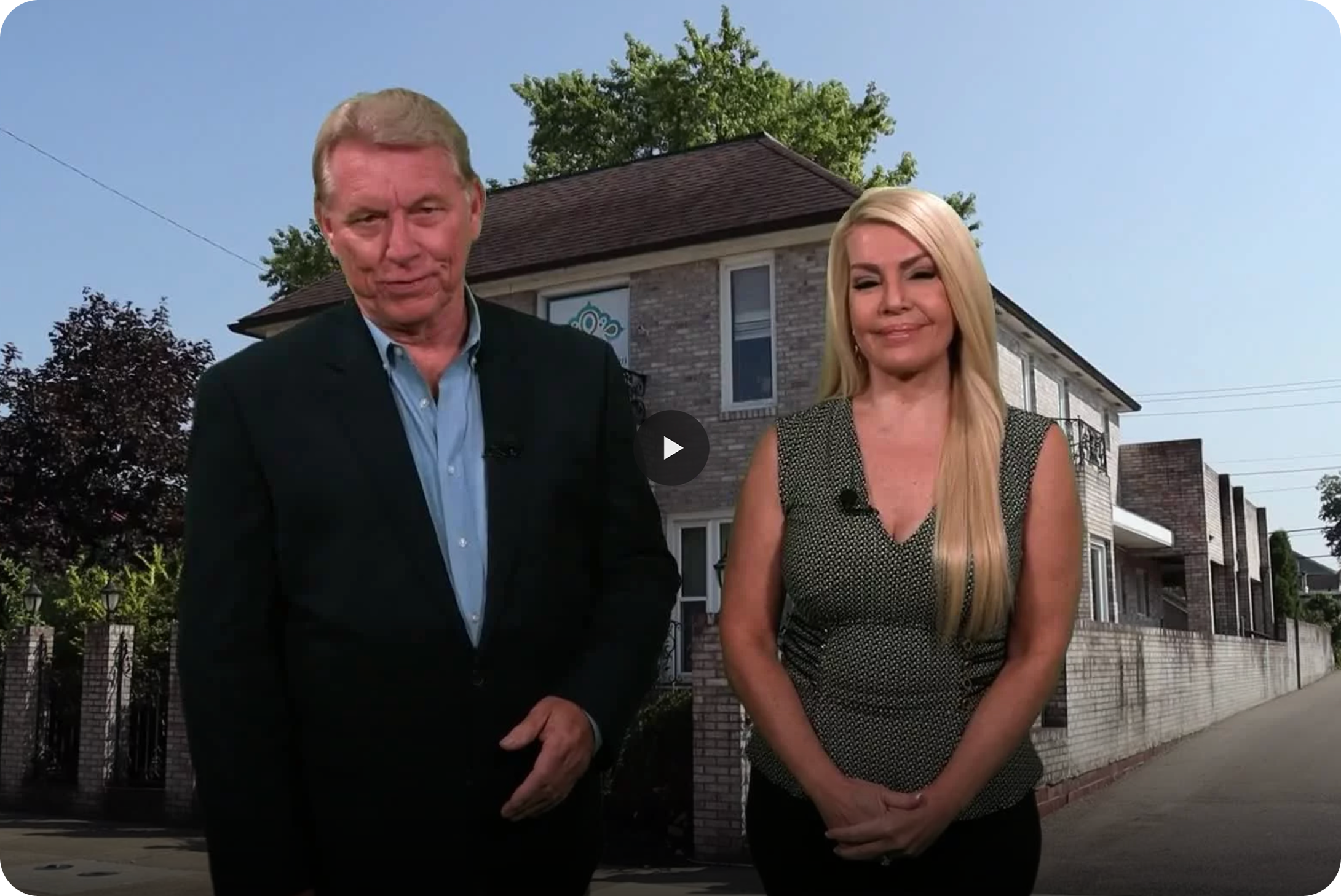 A man and a woman standing outside in front of a house during daytime, both wearing microphones, with relaxed expressions.