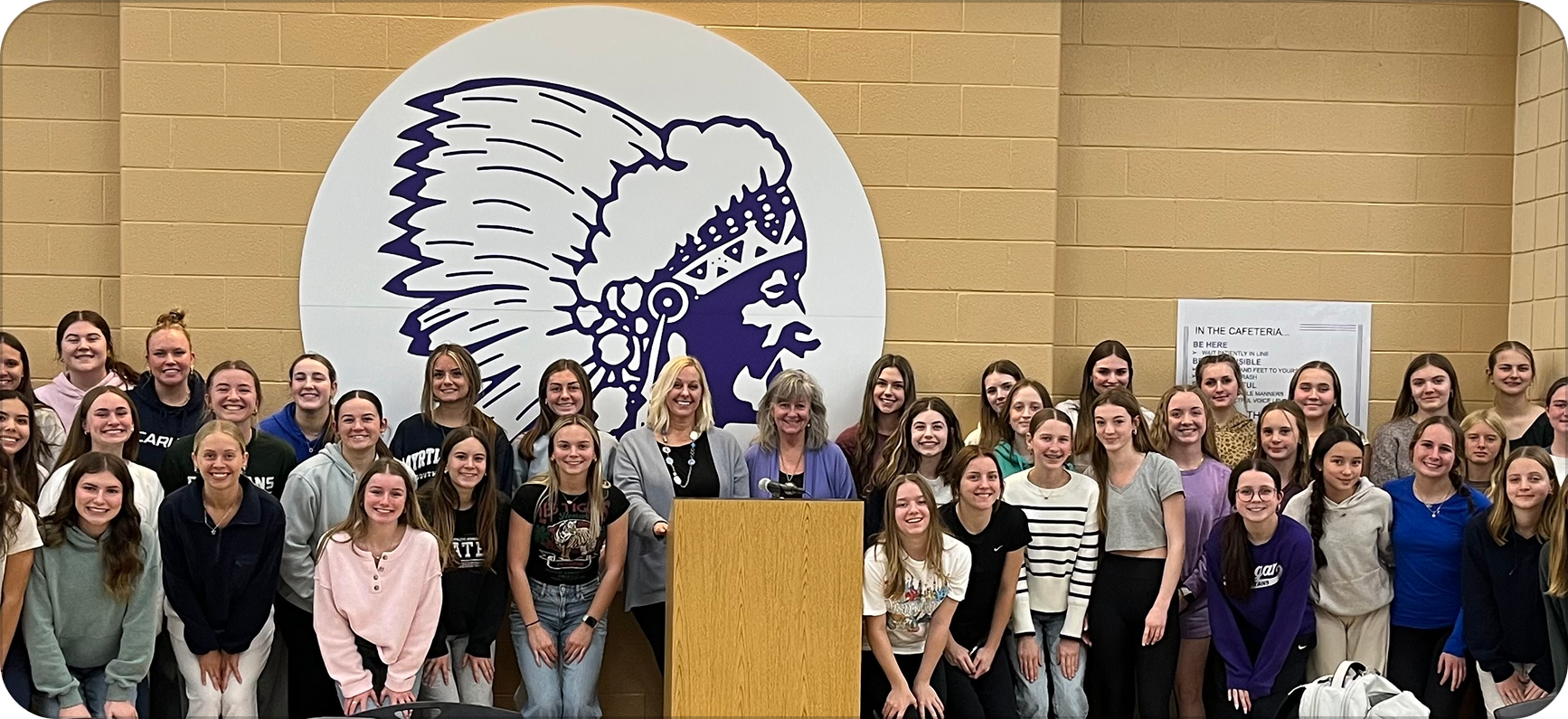 Group of smiling young women and girls gathered around a wooden podium with two women standing behind it in a school hallway, with a large school logo featuring the Logan Chieftan school logo on the yellow brick wall behind them.
