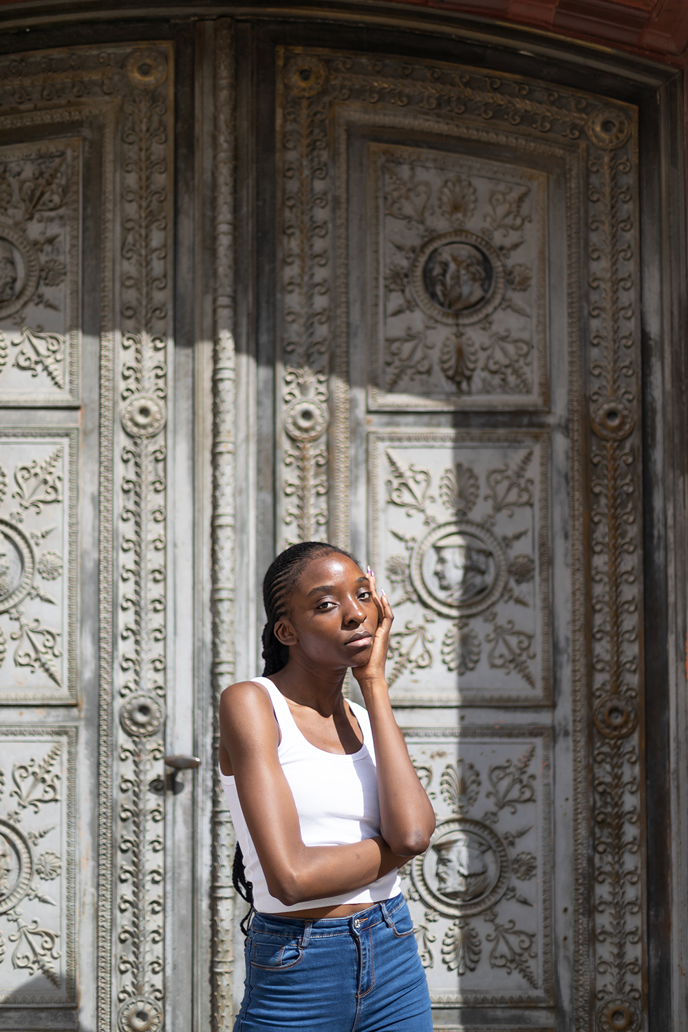 Junge Frau mit gepackten Haaren in weißen Tanktop und blauer Jeans steht vor einer kunstvoll verzierten antiken Tür, die mit floralen und menschlichen Gesichtern verziert ist, im Sonnenlicht.