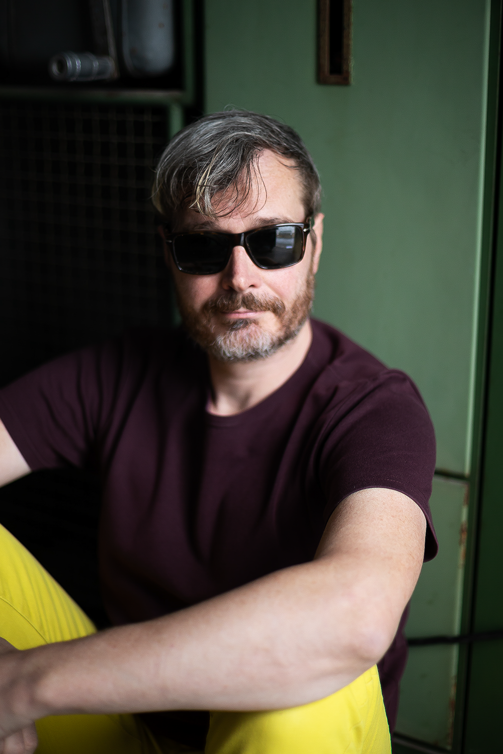 Ein Mann, Dj- mit Sonnenbrille und Bart sitzt in einem Raum mit grünen Schränken im Hintergrund.