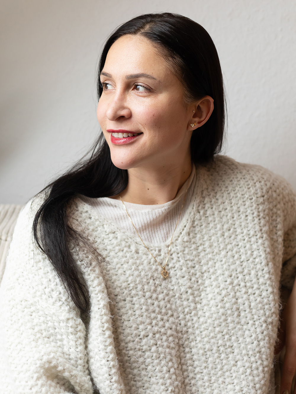 Nahaufnahme einer lächelnden Frau mit schwarzen Haaren, die eine weiße, strukturierte Strickjacke trägt, vor einem hellen Hintergrund.