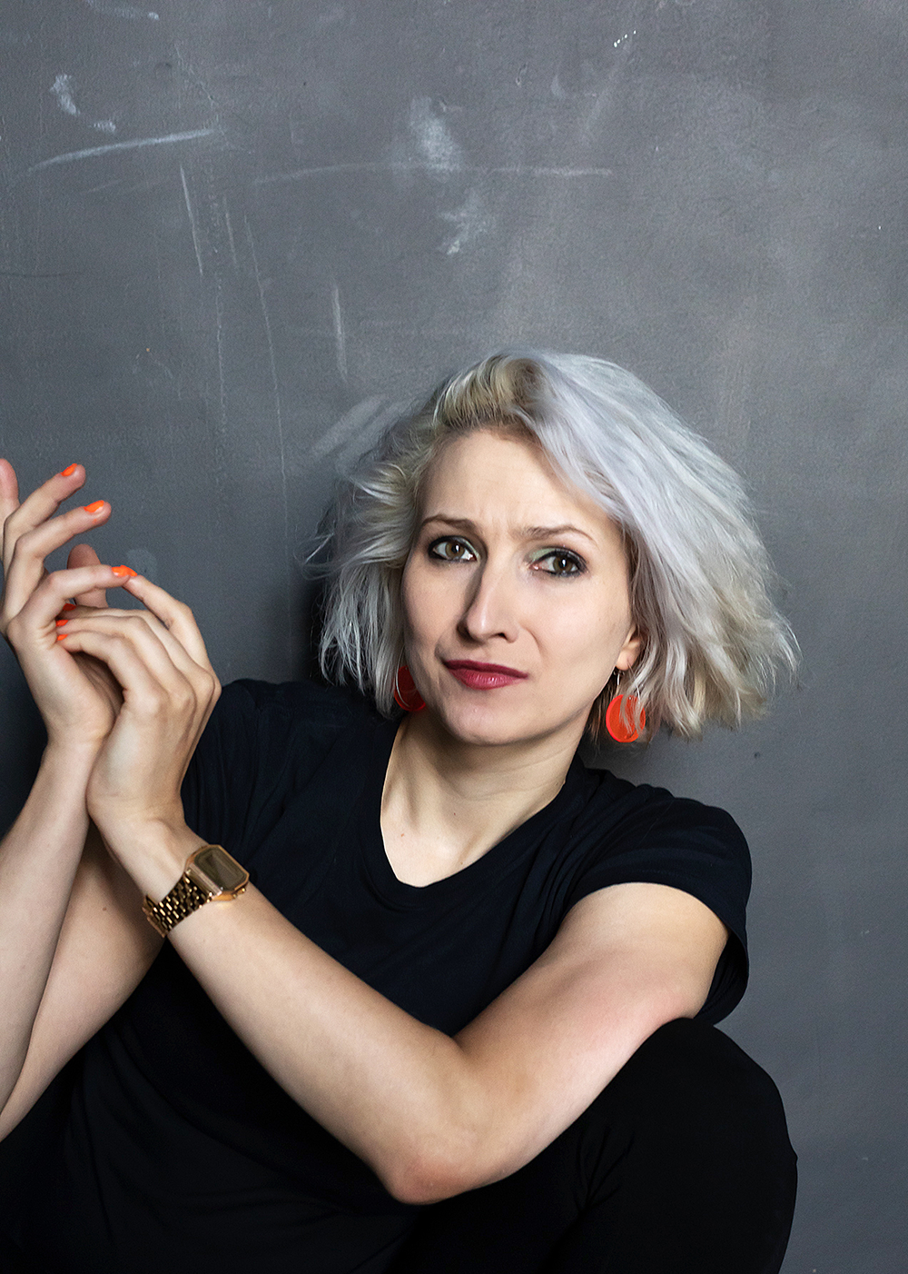 Eine Frau mit blonder, zerzauster Haar, trägt ein schwarzes T-Shirt, rote Ohrringe und eine goldene Armbanduhr, sitzt vor einer grauen Wand, schaut nach vorne mit einem skeptischen Ausdruck.