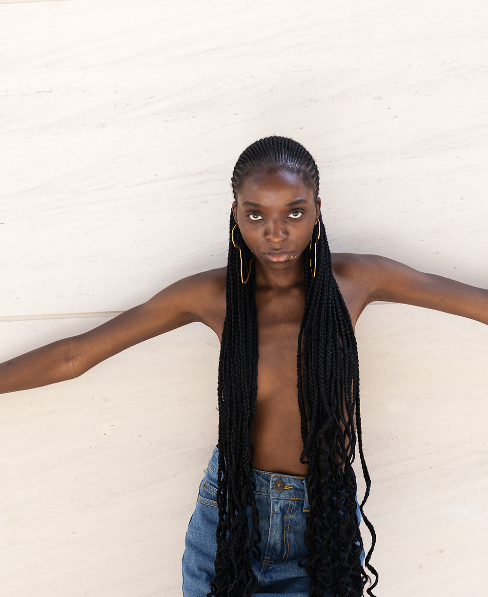 Junge Frau mit langen Zopfhaaren, die nach vorne fallen, posiert mit ausgestreckten Armen vor einer weißen Wand.