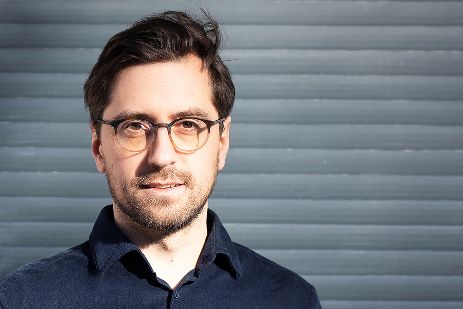 Porträt eines jungen Mannes mit Brille vor einer grauen, gestreiften Wand