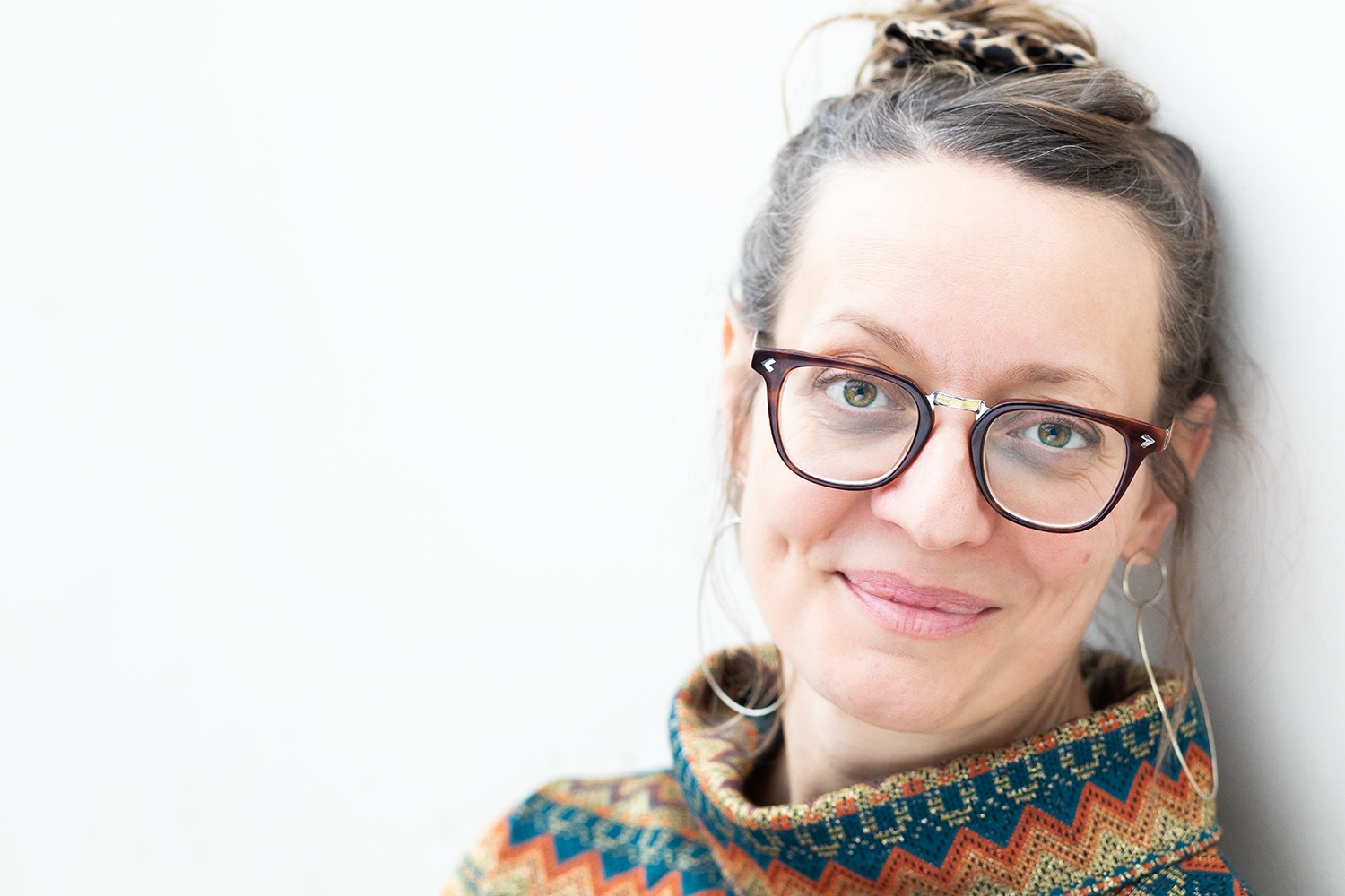 Nahaufnahme eines lächelnden Mädchens mit Brille, Afro-Locken, Sternenohrring und bunter Tuch, lehnend an eine weiße Wand.