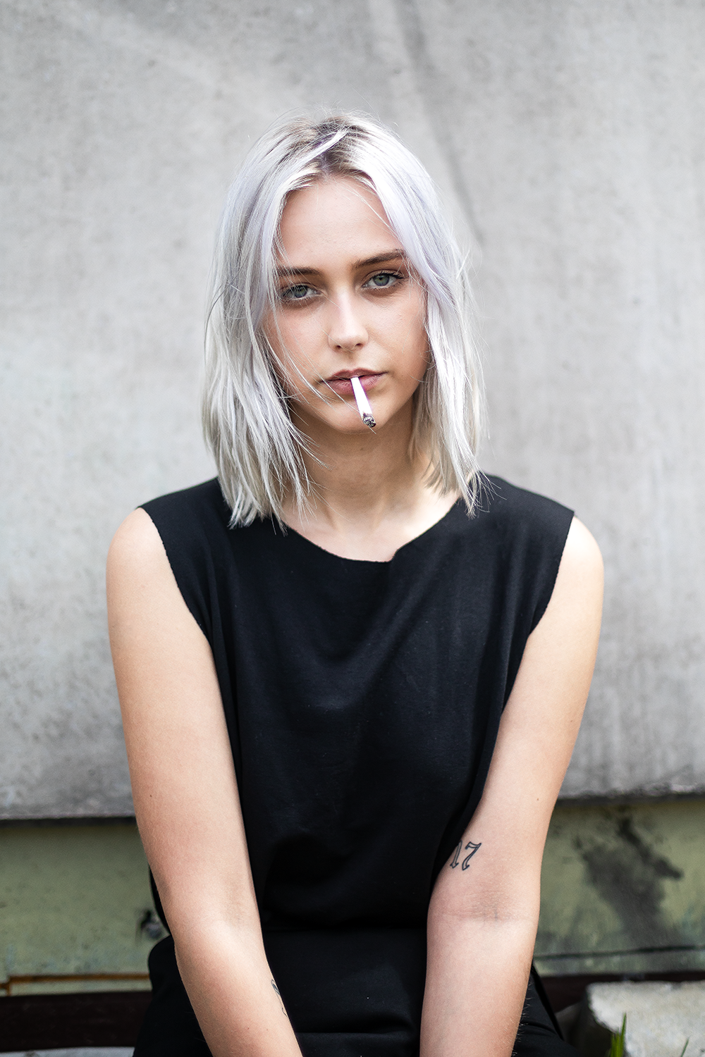 Junge Frau mit hellblonden, zerzausten Haaren trägt ein schwarzes Ärmel-loses Oberteil und raucht eine Zigarette. Sie sitzt vor einer grauen Betonwand.