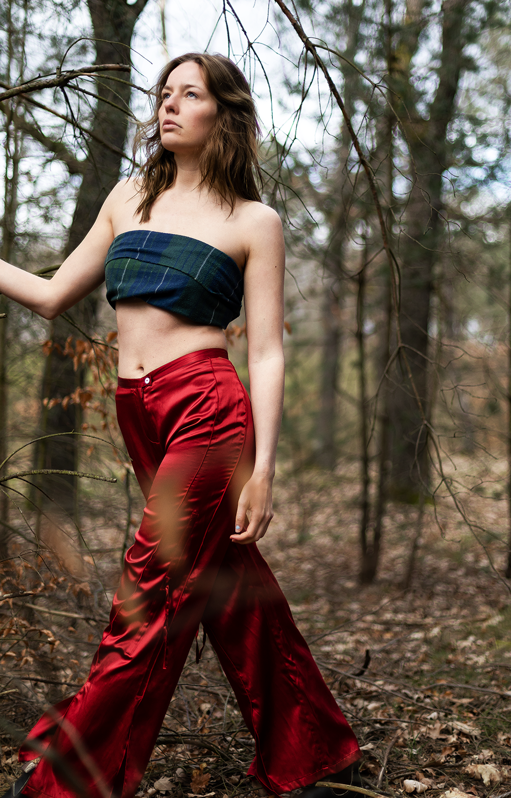 Eine junge Frau in blauer Stoffhose und roten Seidenhosen steht im Wald, umgeben von Bäumen und Laub.