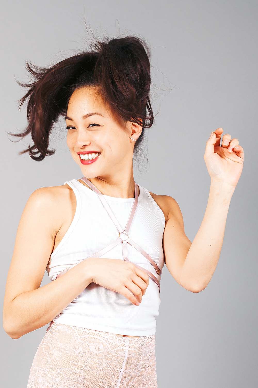 Junge Frau mit dunkelbraunem, voluminösem Haar, smiling, trinkt aus einer Wasserflasche, trägt einen weißen Tanktop und eine durchsichtige Spitzenhose vor grauem Hintergrund.