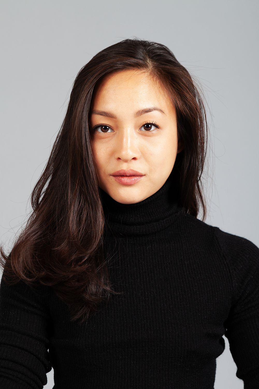 Nahaufnahme eines jungen asiatischen Mädchens mit langen braunen Haaren, die einen schwarzen Rollkragenpullover trägt, vor neutralem grauem Hintergrund.