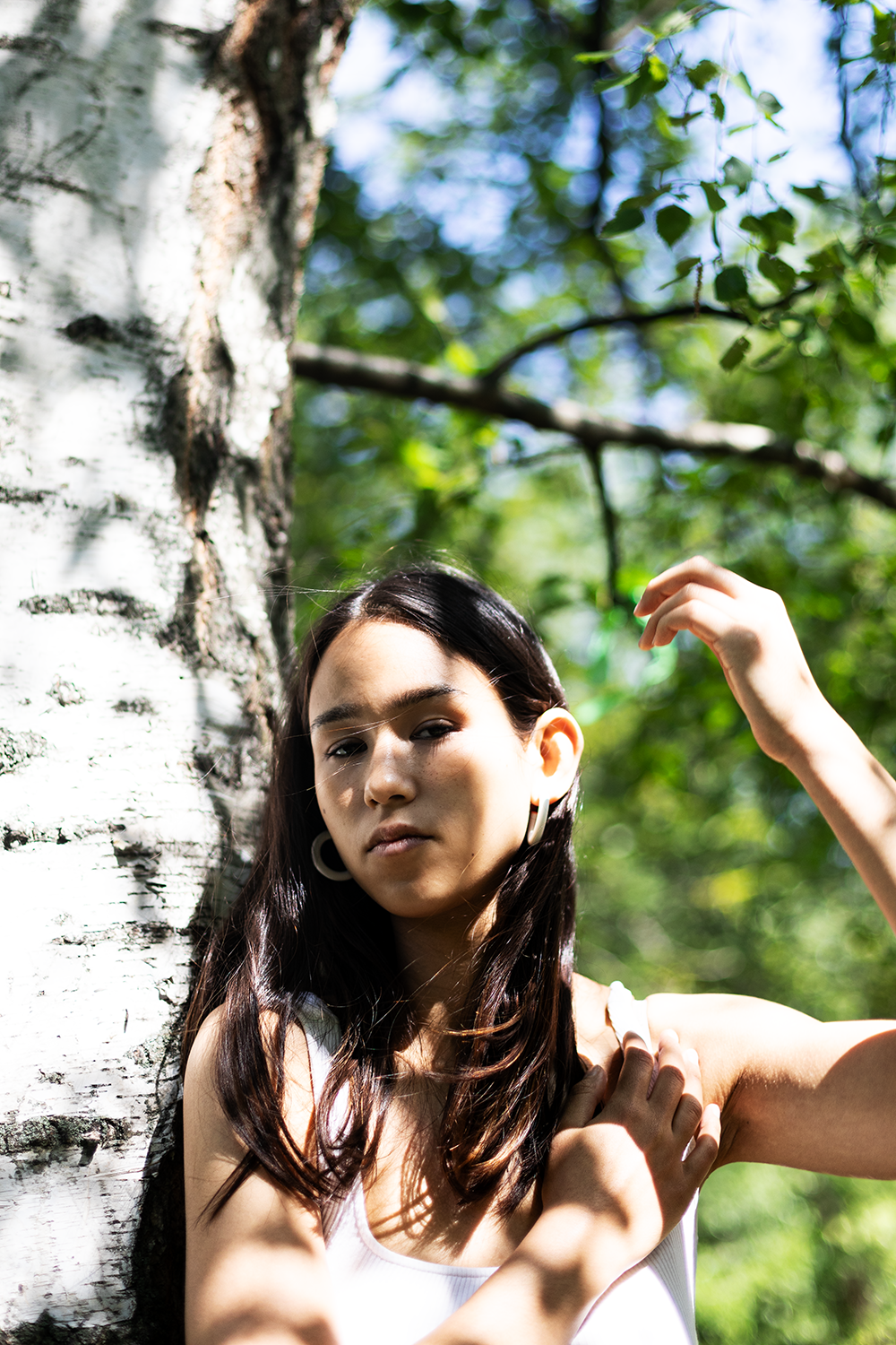 Junge Frau lehnt an einem Baum im Wald, umgeben von grünen Blättern, mit Sonnenlicht, das auf ihr Gesicht fällt.