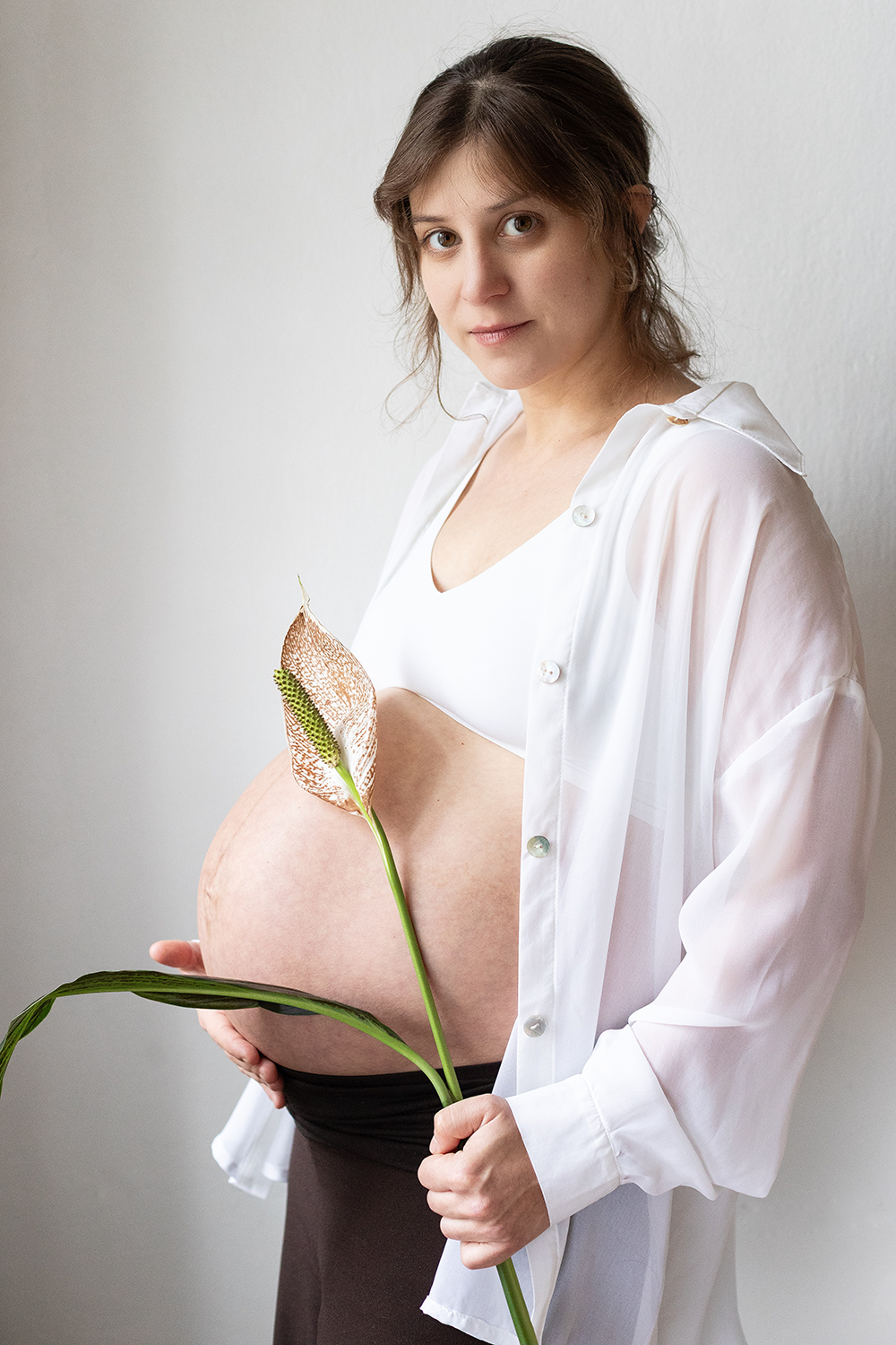 Schwangere Frau mit weißen Kleid und offener Bluse, die eine Calla-Lilie in der Hand hält, vor weißer Wand.