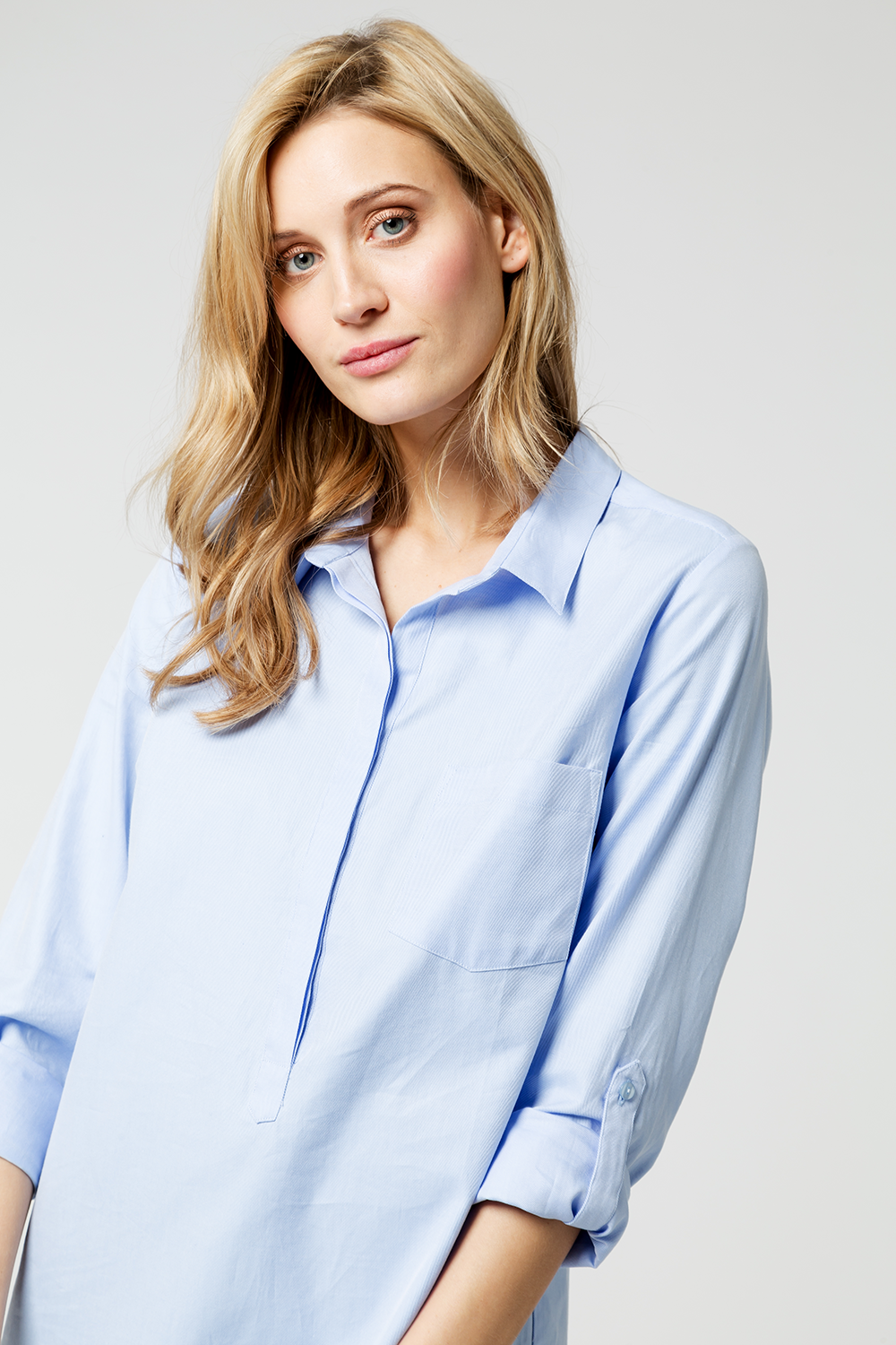 Junge Frau mit blonden Haaren in blauer Hemd, frontal Blick, neutraler Hintergrund.