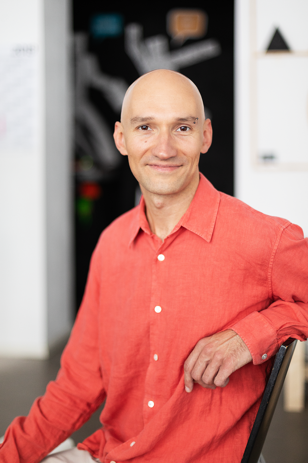 Ein Mann mit Glatze und einem freundlichen Lächeln, der in einem modernen Arbeitsumfeld sitzt, trägt ein rotes Hemd.