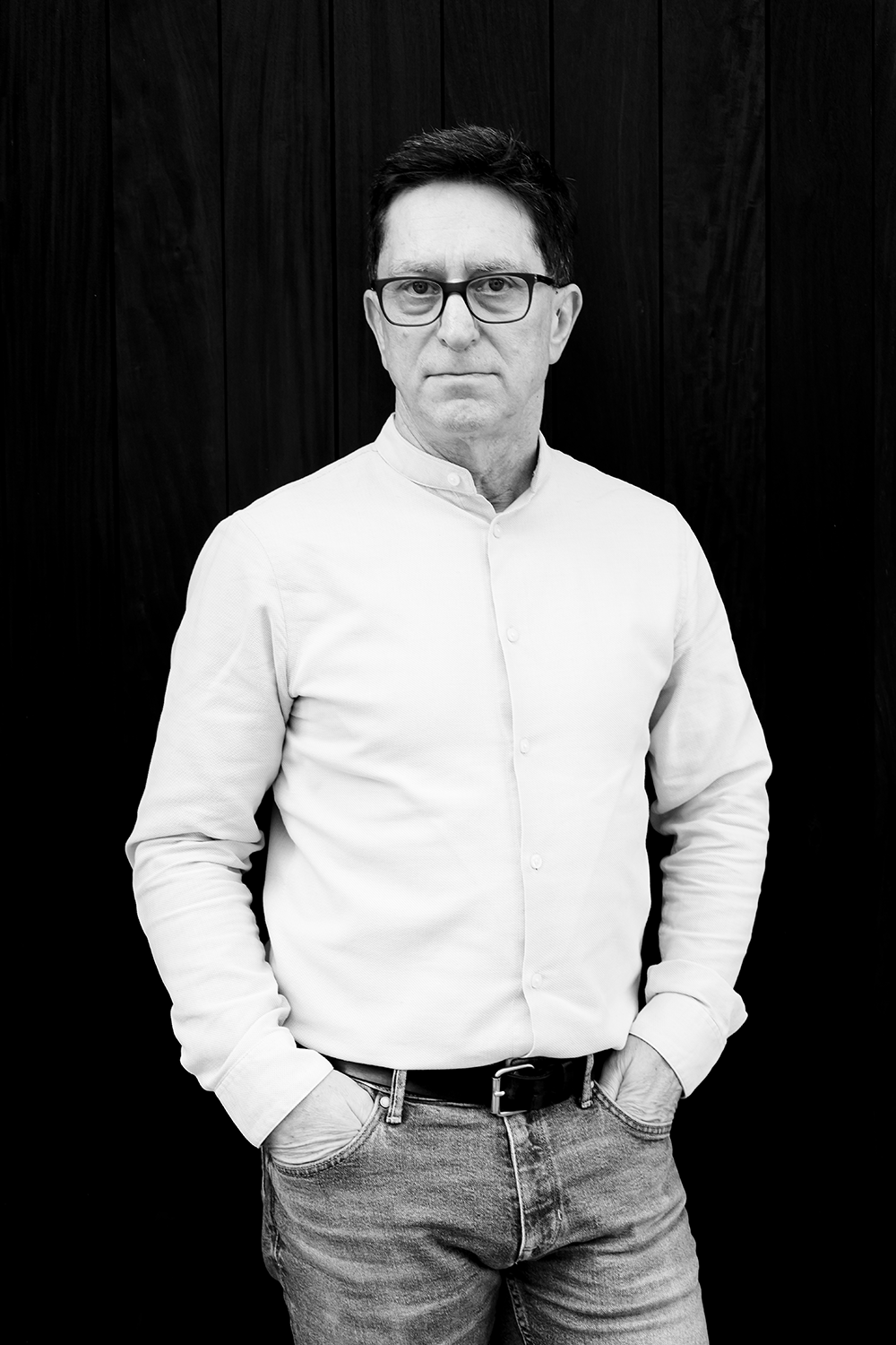 Schwarz-Weiß-Foto eines Mannes mit Brille, weißem Hemd und Jeans, stehend vor einer dunklen Holzwand, mit Händen in den Taschen.