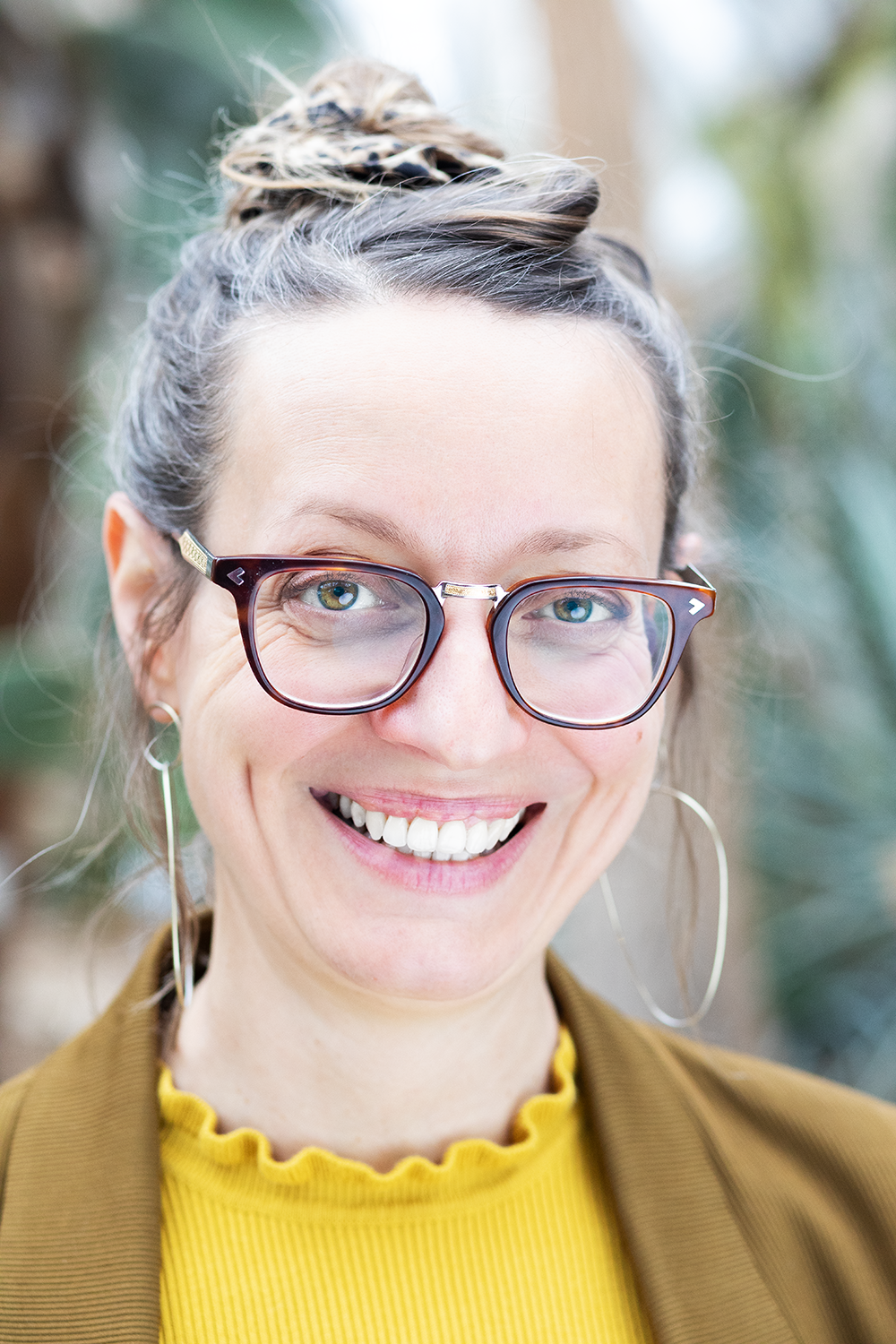 Lächelnde Frau mit Brille, gelbem Oberteil und Ohrringen vor unscharfem grünen Hintergrund.