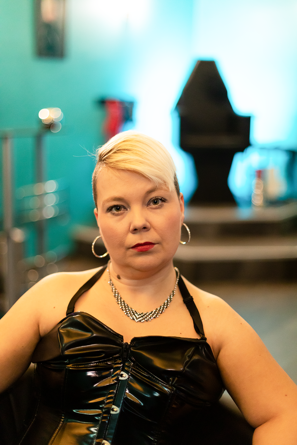 Eine Frau mit blonder, seitlich kurz geschnittener Frisur, trägt ein schwarzes, glänzendes Oberteil und silberne Schmuckstücke, sitzt vor einer blauen beleuchteten Klavier im Hintergrund.