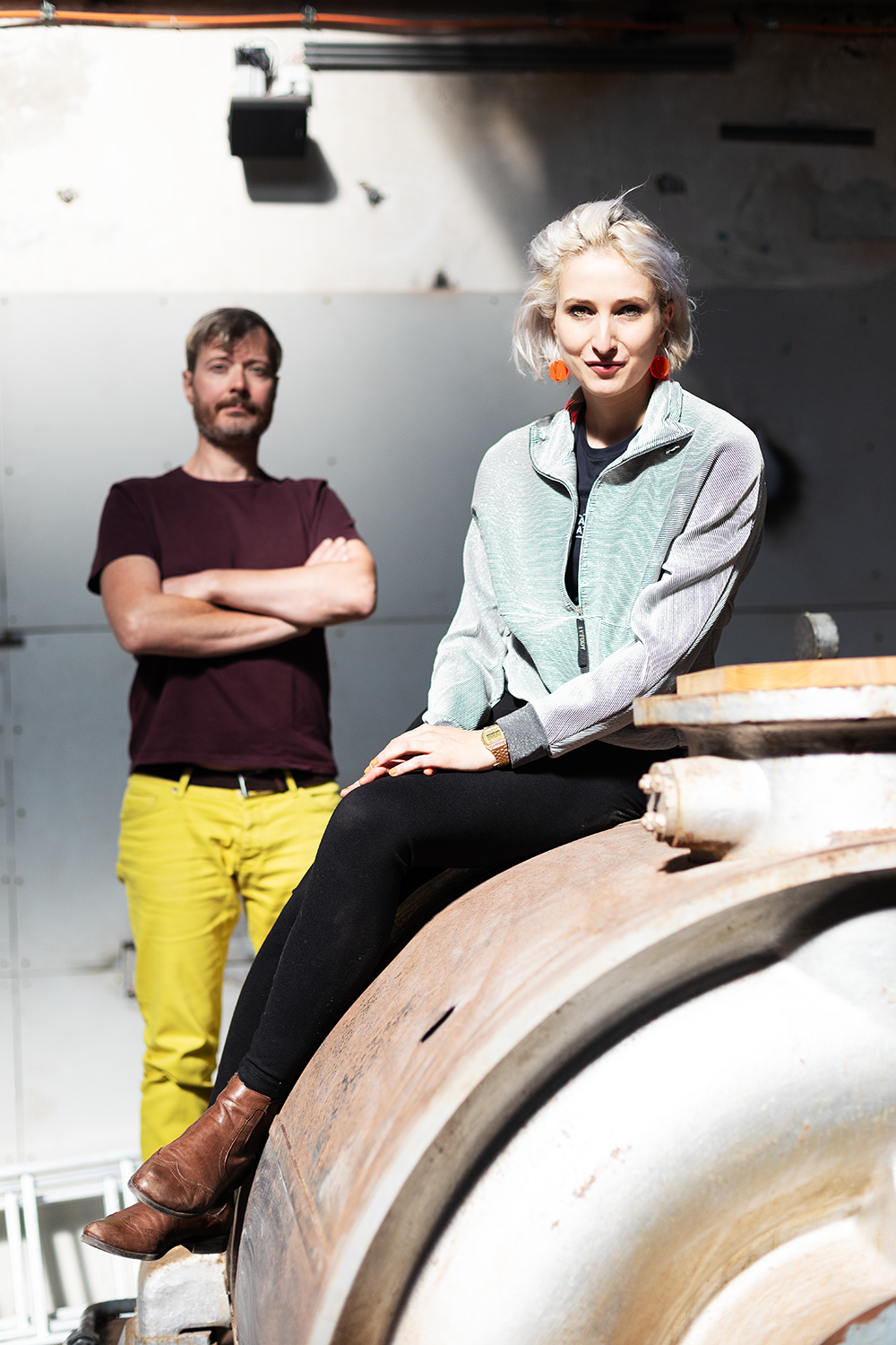 Eine Frau mit blonder, lockiger Frisur sitzt auf einem großen, alten Metallzylinder, während ein Mann mit dunklem Haar und Bart im Hintergrund steht und die Arme verschränkt. Künsterlduo.