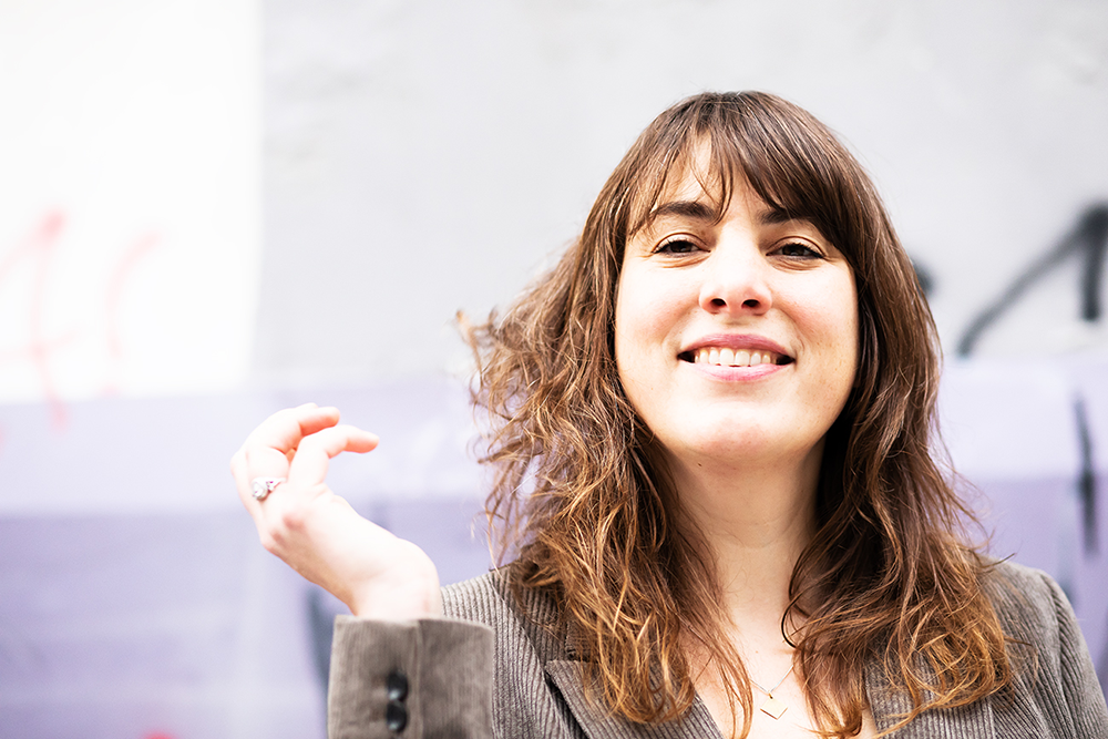 Junge Frau mit braunen, lockigen Haaren, lächelnd, in einem grauen Blazer, vor einer Wand mit Graffiti.
