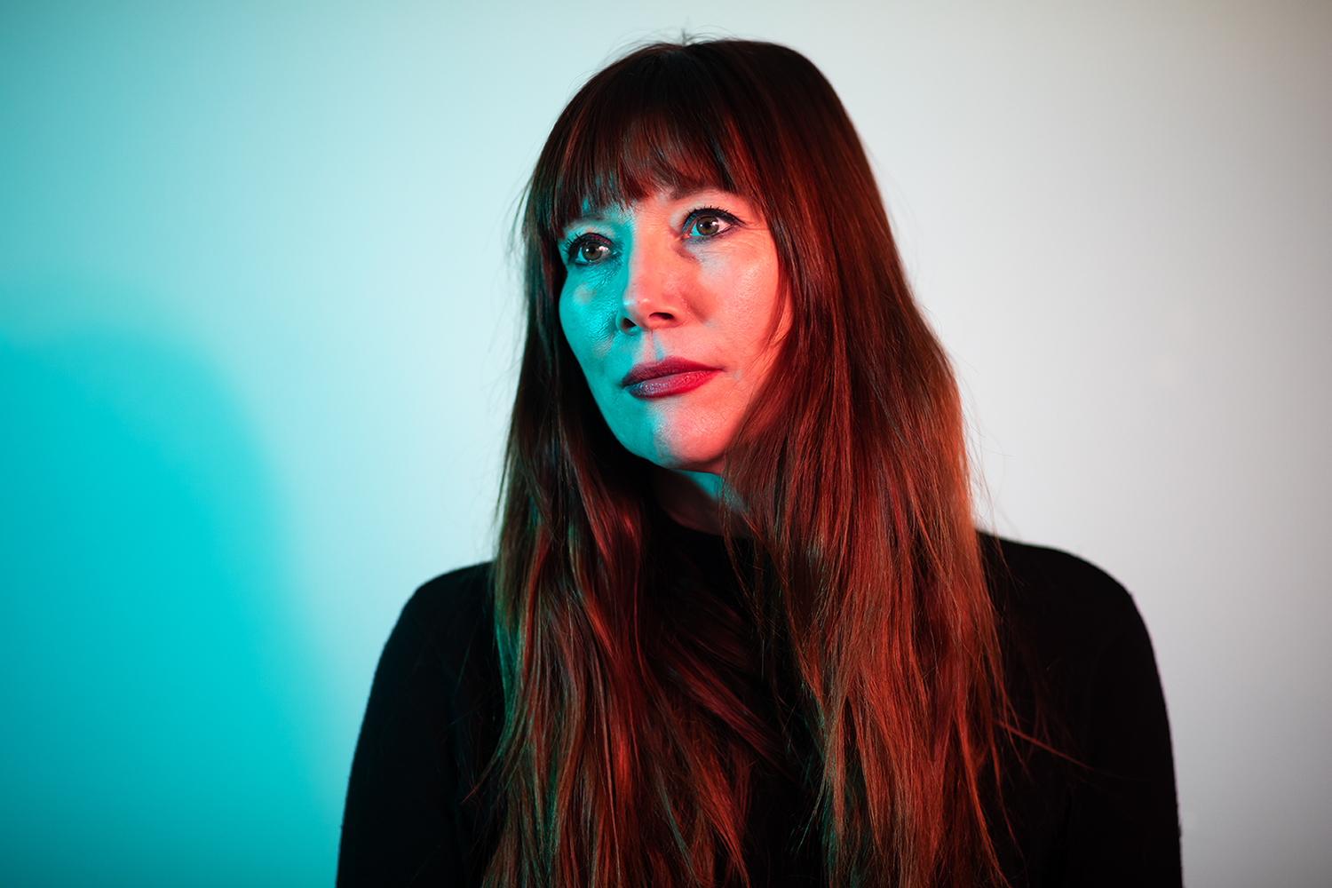 Porträt einer Frau mit langem braunem Haar, schwarze Kleidung, Blick leicht zur Seite, farbige Beleuchtung auf der linken Seite, neutraler Hintergrund.