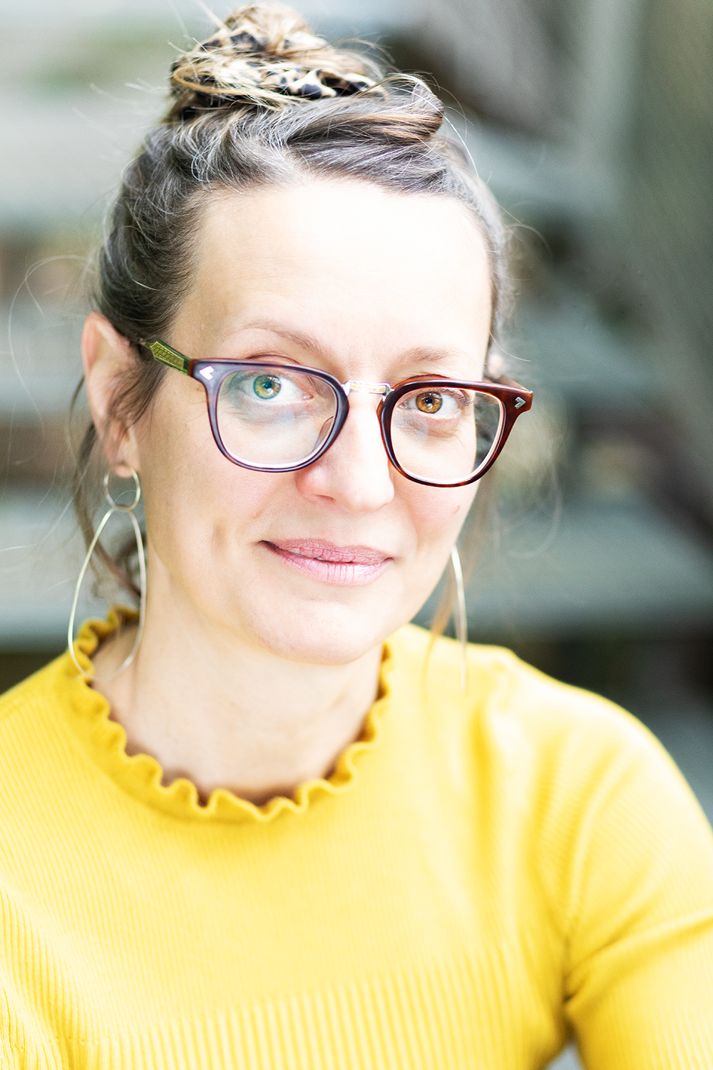 Nahaufnahme einer Frau mit großen Brillengläsern, die lächelnd in die Kamera schaut, trägt eine gelbe Bluse mit Rüschen am Hals, hat lila und grüne Glitzerohrringe, ihre Haare sind hochgesteckt mit einem Leoparden-Print-Etui, im Hintergrund ist ein u