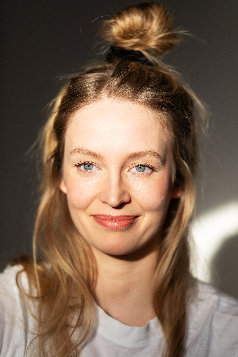 Junge Frau mit blonden, langen Haaren, ist vom Licht beleuchtet, trägt weißen Oberteil, hat einen lockeren Dutt auf dem Kopf, lächelt leicht, Hintergrund dunkel.