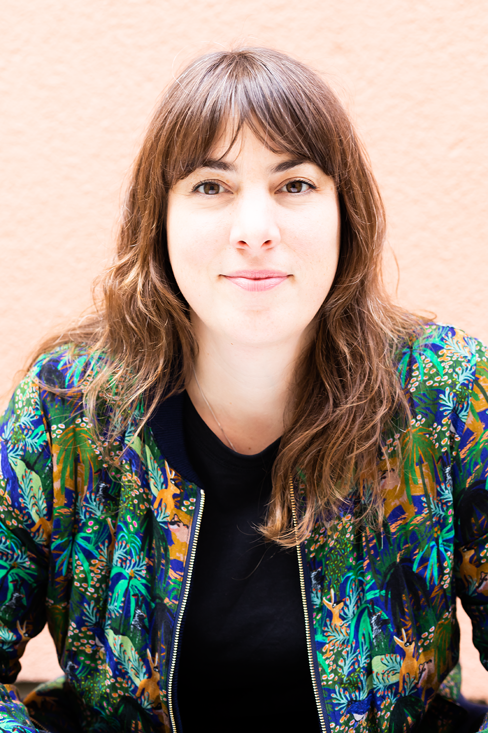 Junge Frau mit braunen, lockigen Haaren trägt ein schwarzes T-Shirt und eine bunte, tropisch bedruckte Jacke, sitzt vor einer hellen Wand.