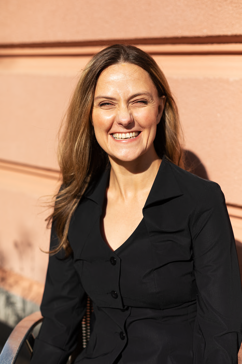 Porträt einer lachenden Frau mit braunen Haaren vor einer sandfarbenen Wand.