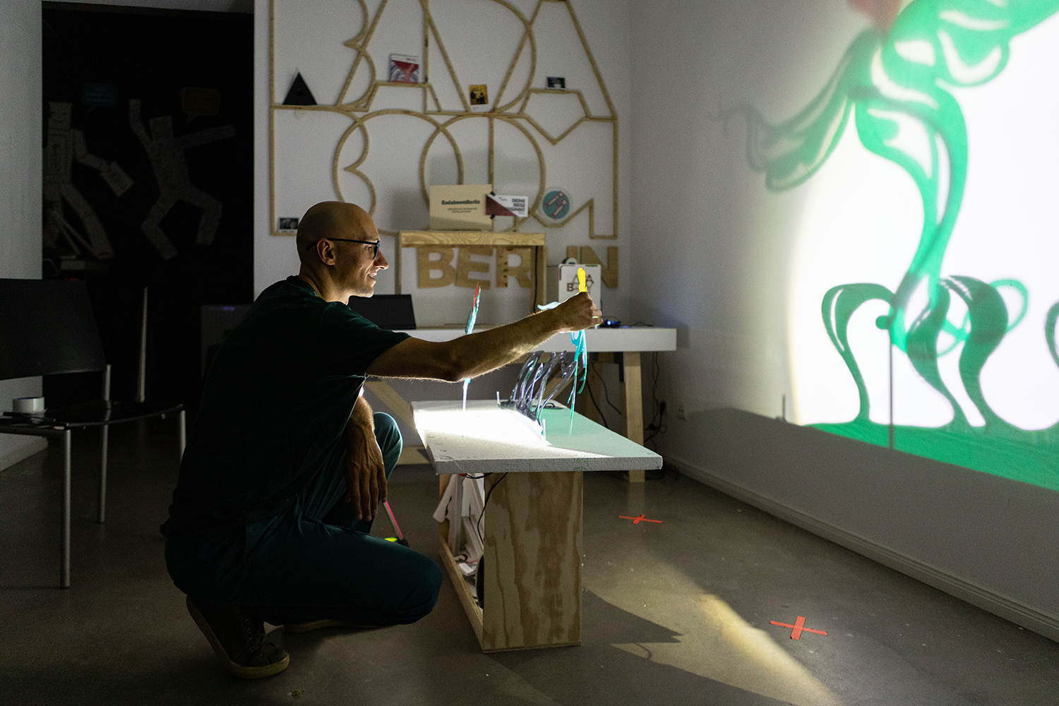 Ein Mann sitzt in einem dunklen Raum vor einer Projektionswand und streckt die Hand aus, während bunte Lichtsträhnen auf ihn und die Wand projiziert werden. Im Hintergrund befinden sich dekorative Regale an der Wand.