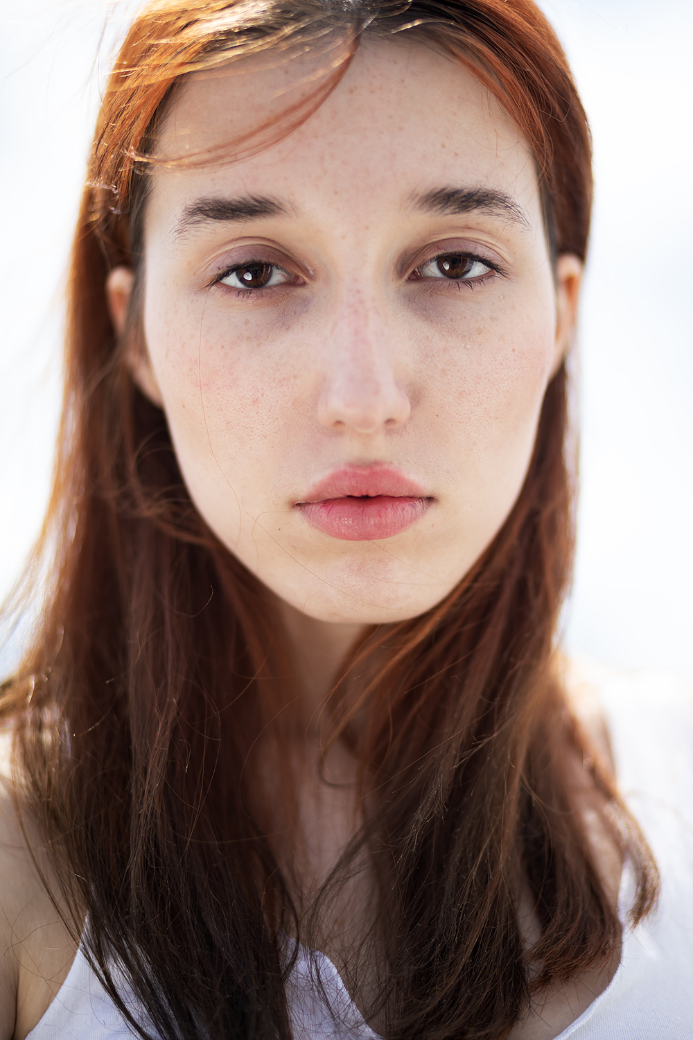 Nahaufnahme eines jungen Mädchens mit langen braunen Haaren, Sommersprossen, braunen Augen und natürlichen Lippen, leichtem Blick und hellem Hintergrund.