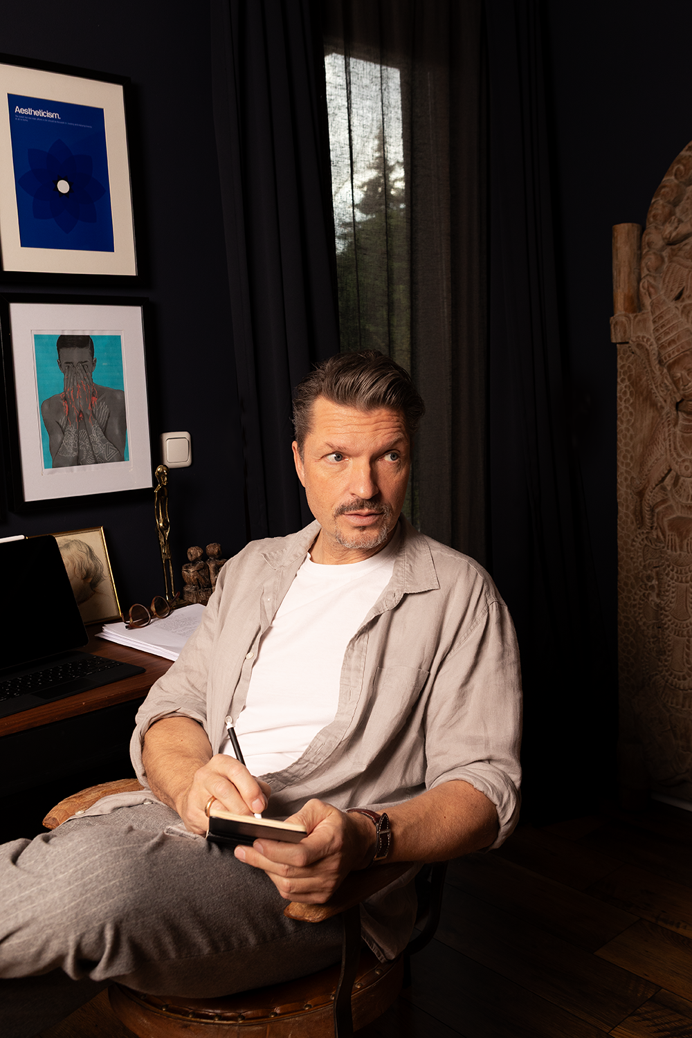 Ein Mann sitzt in einem modernen, dunklen Raum, schreibt in ein Notizbuch und schaut nachdenklich zur Seite. Im Hintergrund sind Kunstwerke an der Wand, ein Laptop und einige Dekorationsgegenstände auf einem Schreibtisch.