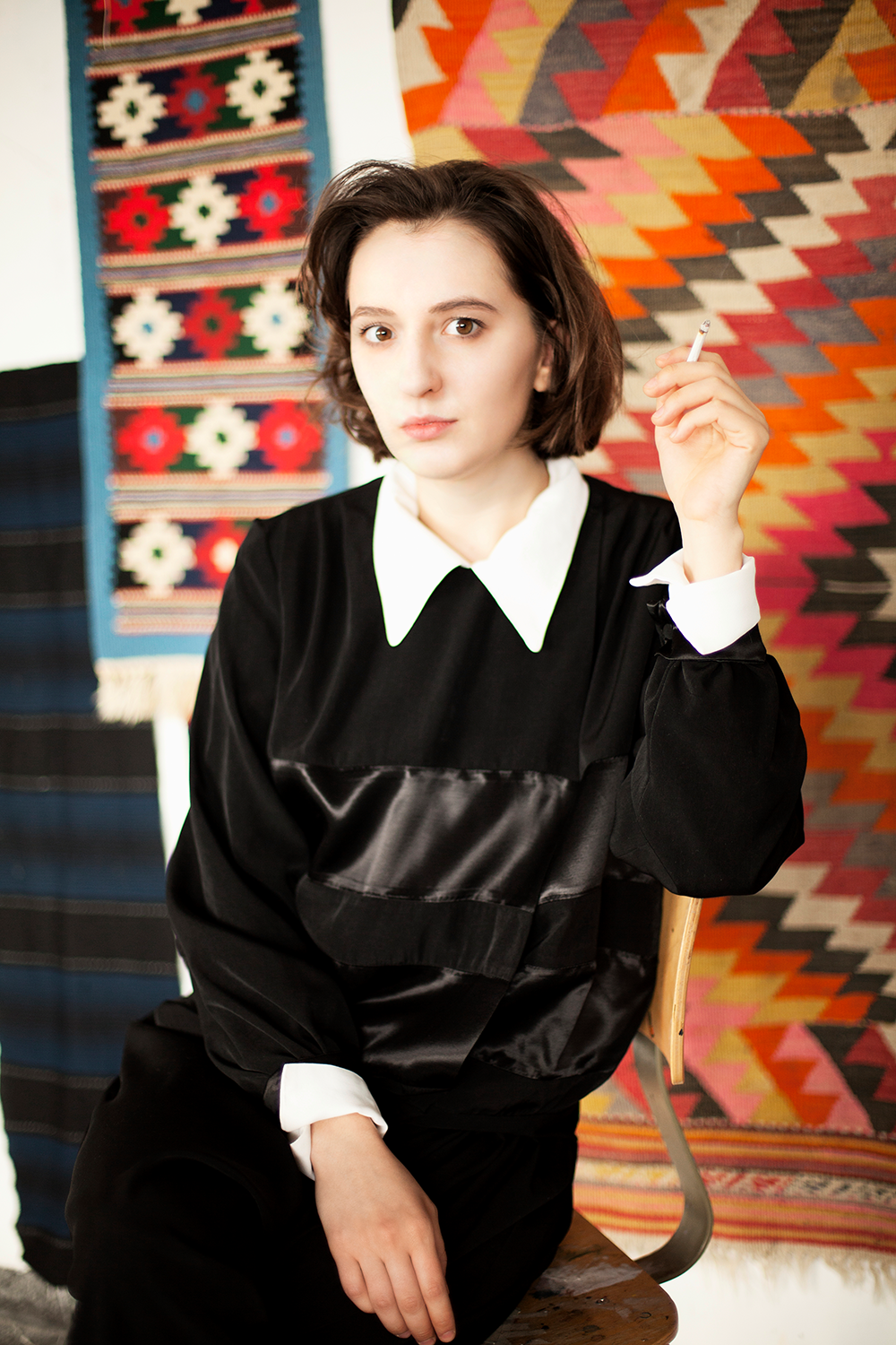 Junge Frau mit dunklem Kleid mit weißen Kragen und Manschetten, sitzend vor bunten, gewebten Wandteppichen mit geometrischen Mustern.