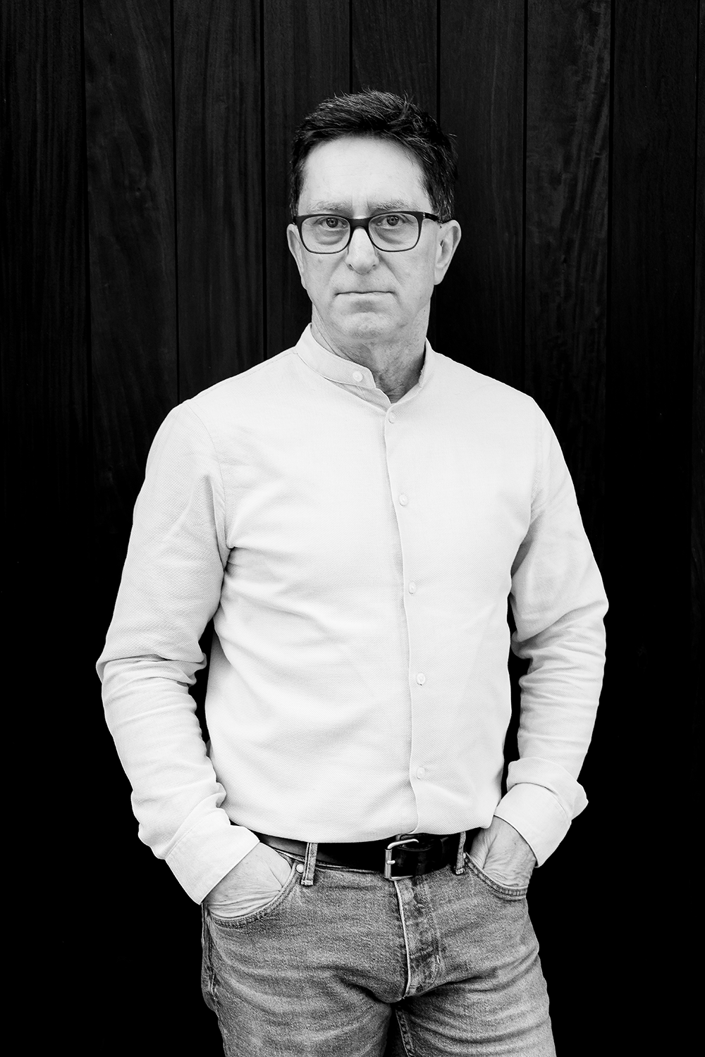 Schwarz-weiß Porträt eines Mannes mit Brille, weißem Hemd, Jeans, stehen vor einer dunklen Holzpaneelwand.
