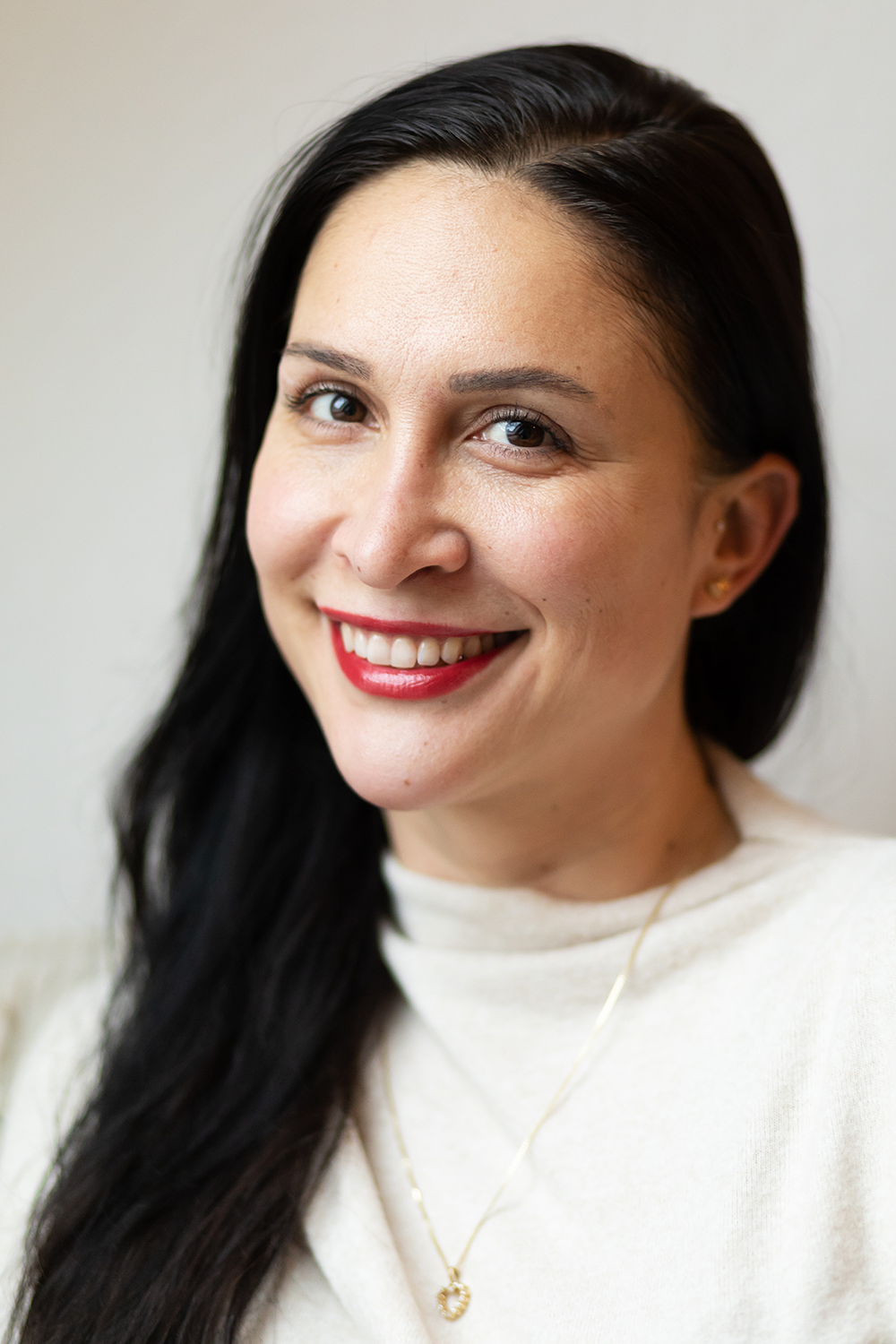 Nahaufnahme einer lächelnden Frau mit dunklem Haar, rotem Lippenstift und goldener Halskette.