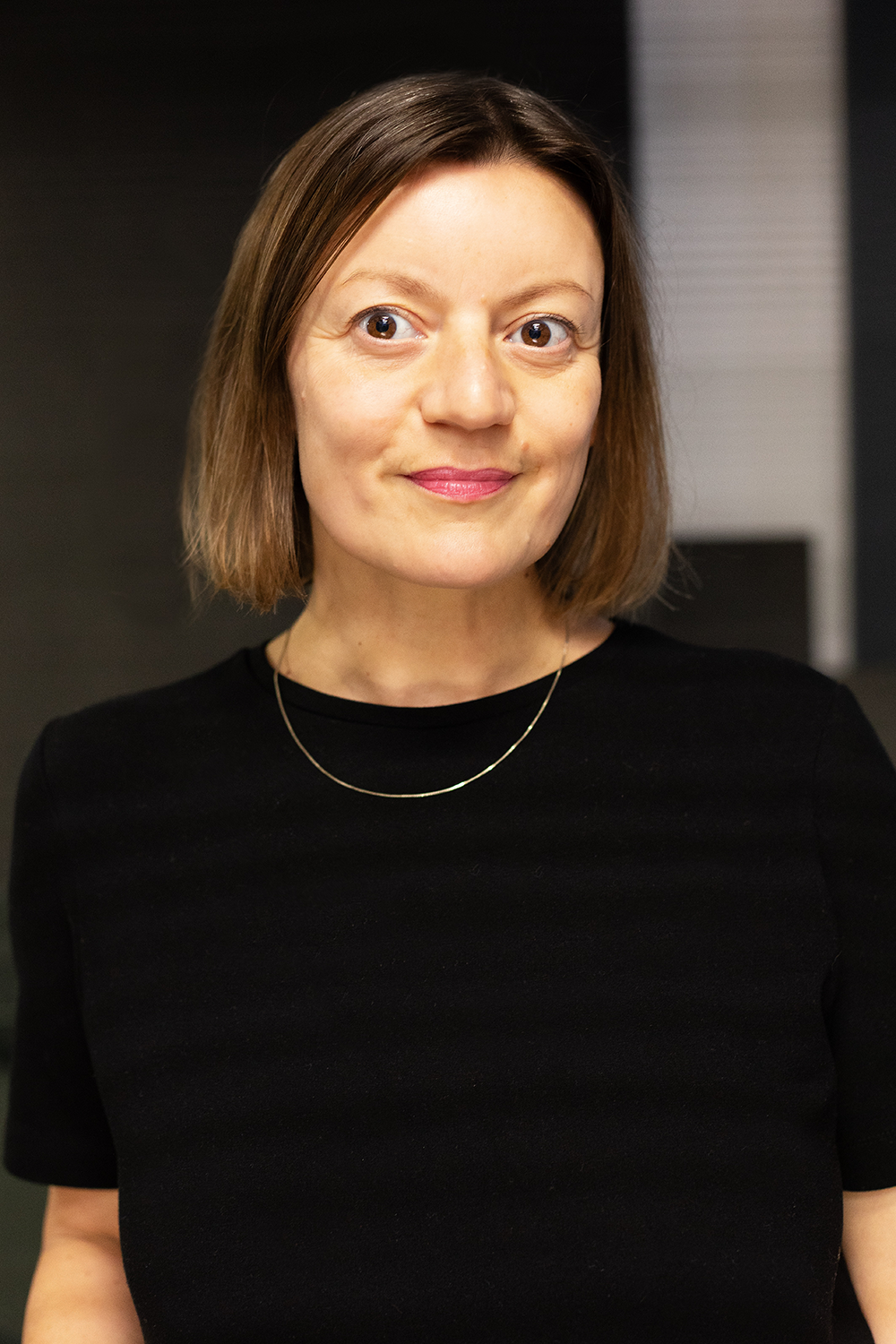 Nahaufnahme einer jungen Frau mit kinnlangen braunen Haaren, die schwarze Kleidung und eine dünne goldene Halskette trägt, vor einem dunklen Hintergrund.