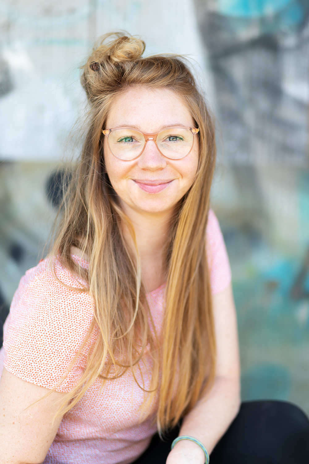 Ein Lächeln eine junge Frau mit roten Haaren, Brille und einem pinkfarbenen Oberteil, sitzend vor einem bunten Hintergrund.