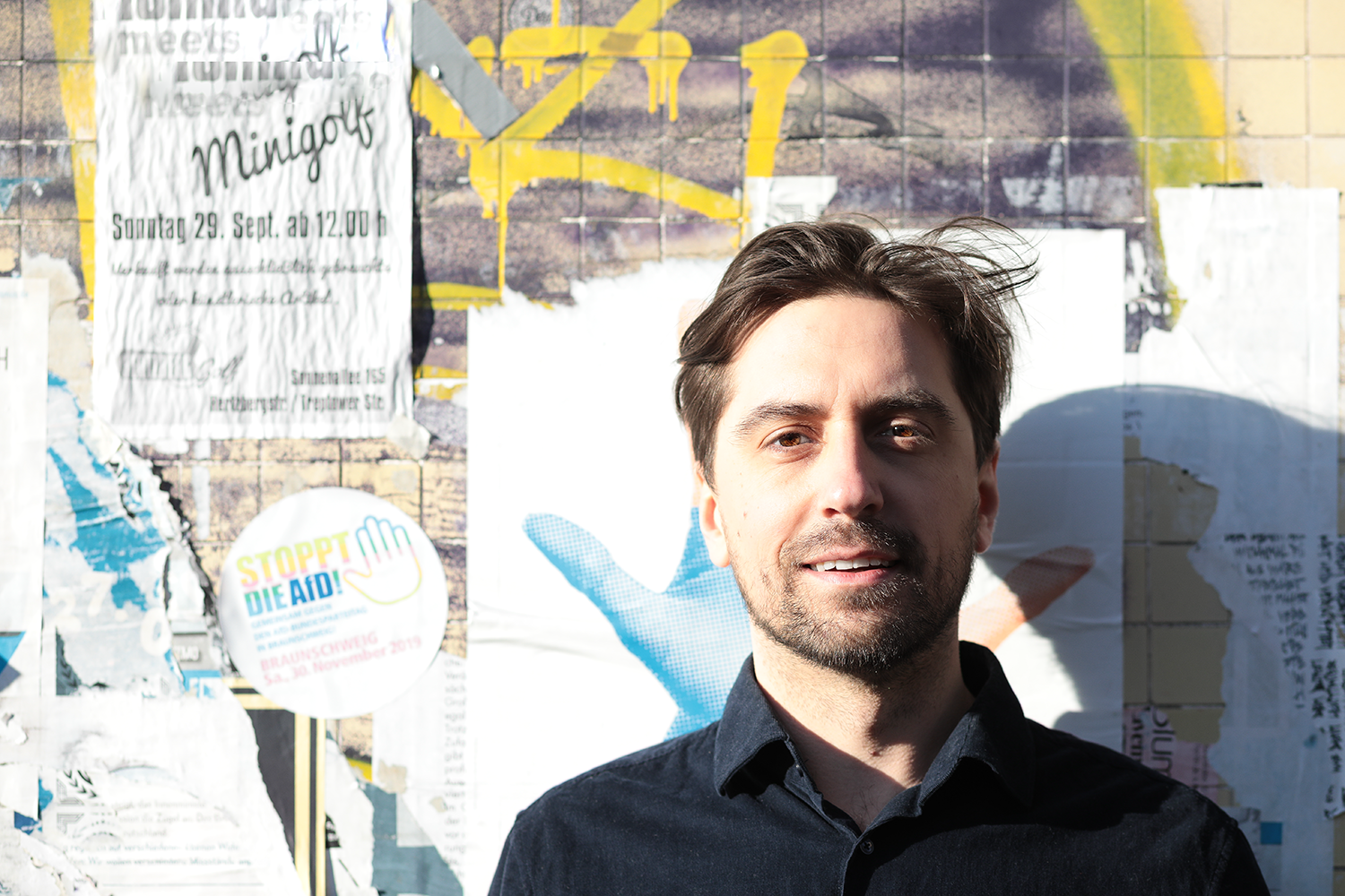 Ein Mann mit dunklem Hemd steht vor einer Wand mit verschiedenen Posters und Flyern, die Werbung und Hinweise enthalten.