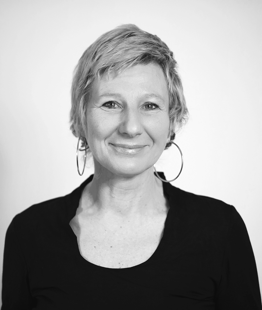 Schwarz-weiß-Porträt einer Frau mit kurzen, lockigen Haaren, großer Ohrringe und schwarzem Oberteil, lächelnd, vor hellem Hintergrund.