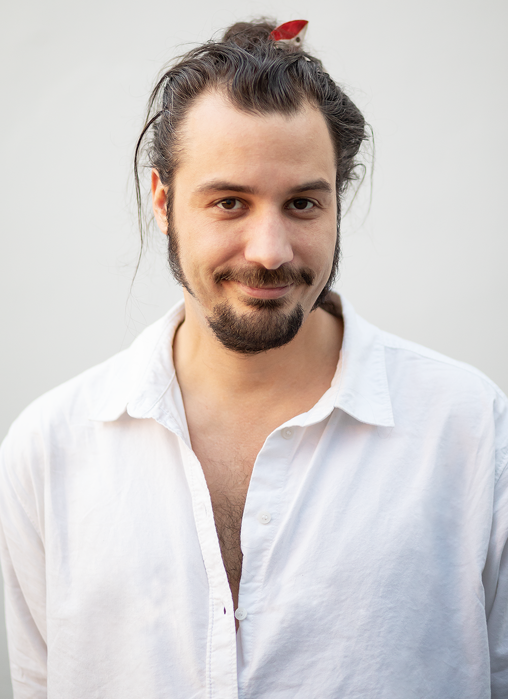 Junger Mann mit langen Haaren, Bart und Schnurrbart, trägt weiße Hemd, vor neutralem Hintergrund.