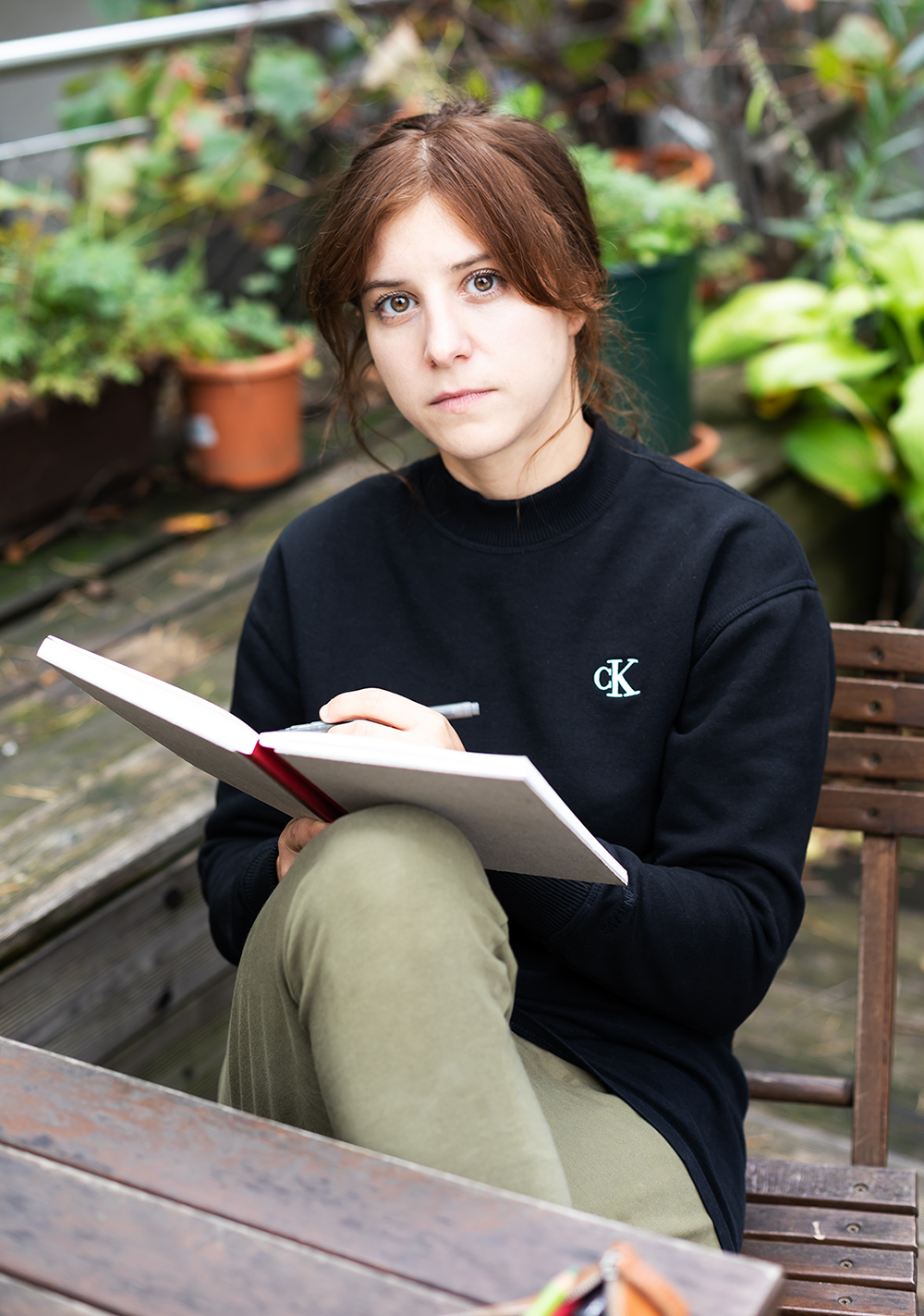 Ein junger Mensch mit braunen Haaren sitzt auf einer Bank im Garten, hält ein Notizbuch und einen Stift, trägt ein schwarzes Sweatshirt mit CK-Logo, umgeben von grünen Pflanzen.