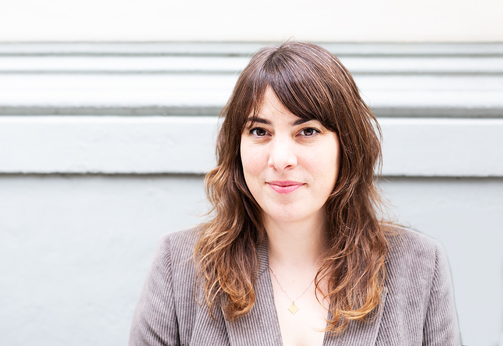 Porträt einer jungen Frau mit braunen, lockigen Haaren, die vor einer weißen Wand steht.