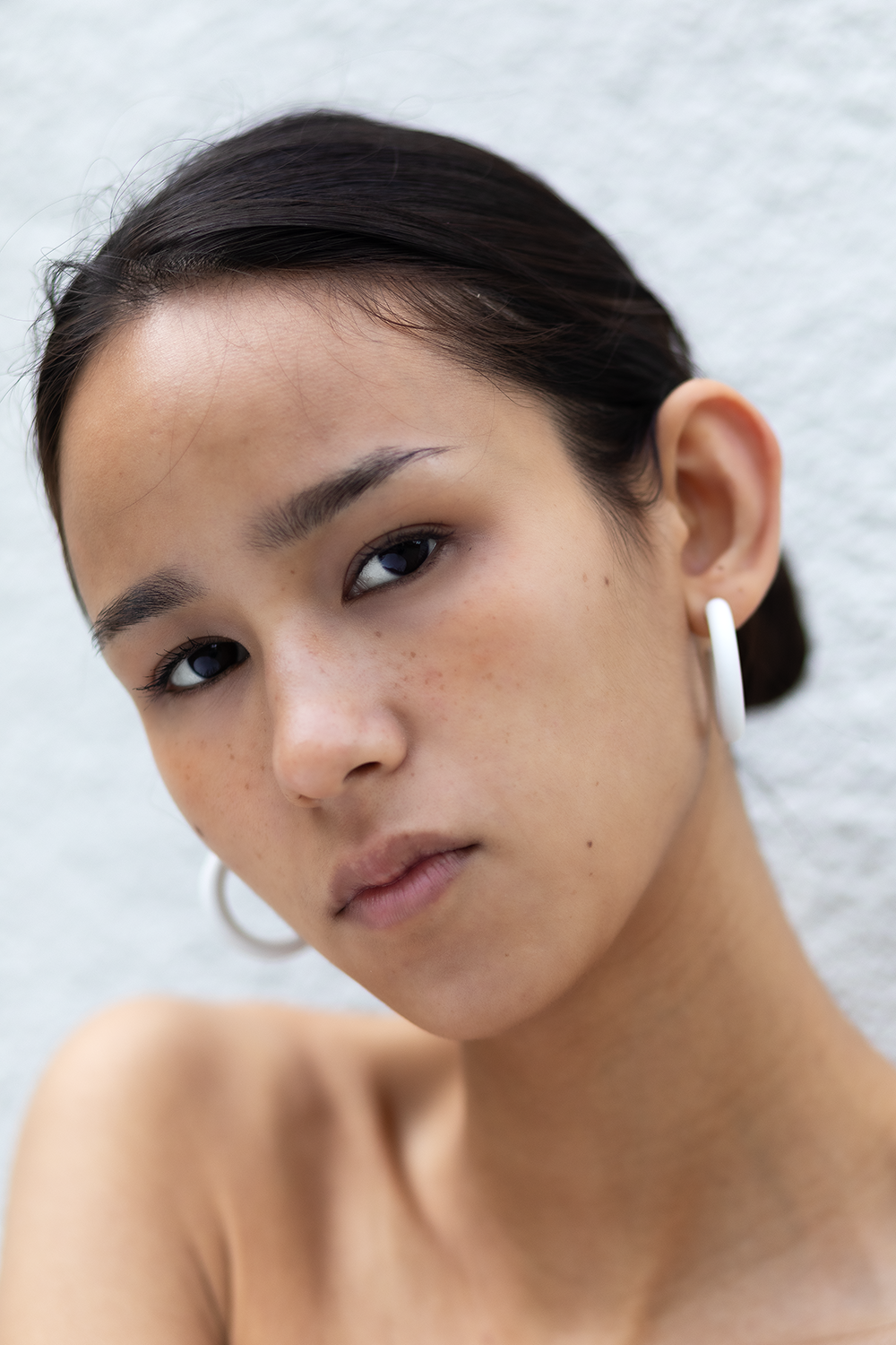 Nahaufnahme eines jungen asiatischen Mädchens mit dunklem Haar, hellbrauner Haut und dunklen Augen, trägt kreisförmige weiße Ohrringe, vor hellem grauem Hintergrund.