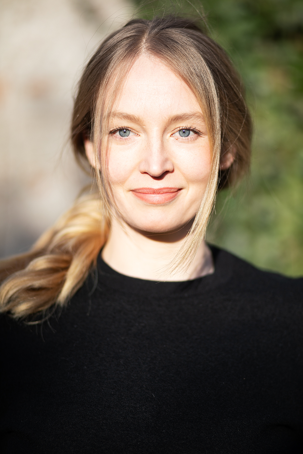 Nahaufnahme eines lachenden jungen Mädchens mit blonden, lockigen Haaren vor einem grünen und hellen Hintergrund.