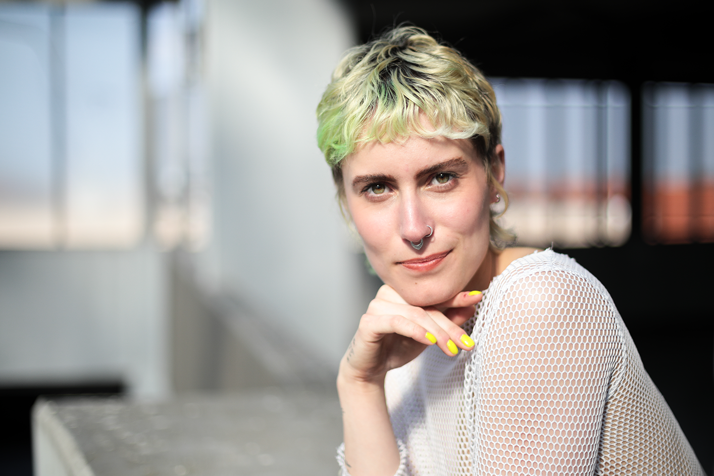 Junge Frau mit kurzen, blonden und grünen Haaren trägt ein weißes Netz-Shirt, hat einen Nasen- und einen Septum-Piercing, trägt gelbe Fingernägel und lächelt in die Kamera.