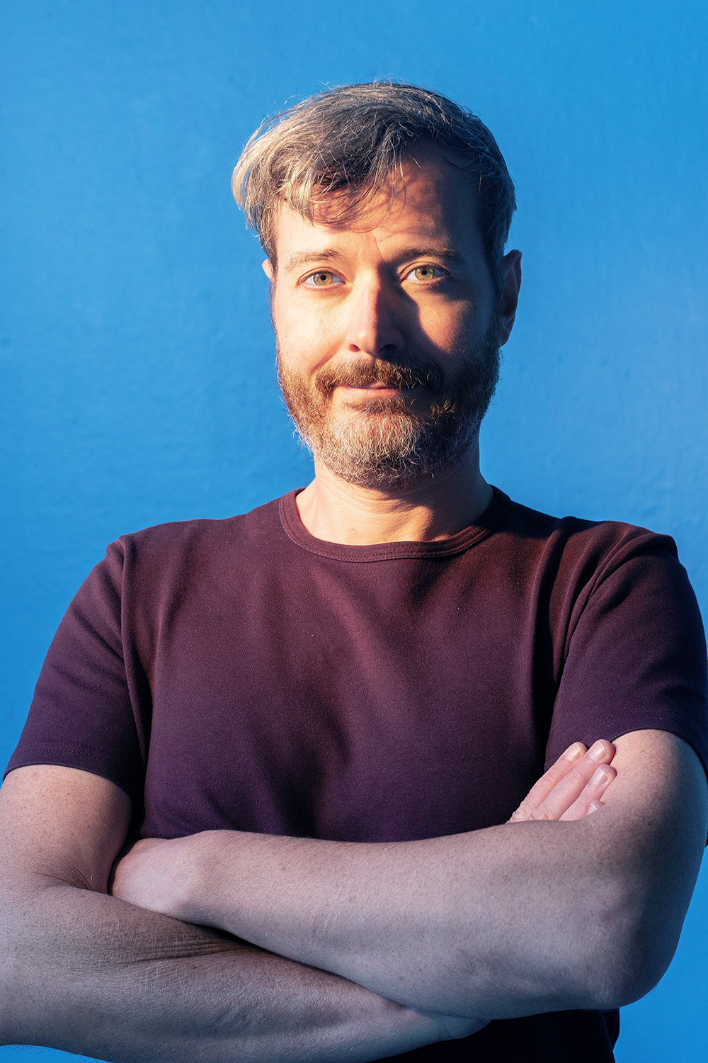 Ein Mann mit einem Bart und blonden Haaren, trägt ein dunkelrotes T-Shirt, steht vor einem blauen Hintergrund, wirkt selbstbewusst und entspannt.