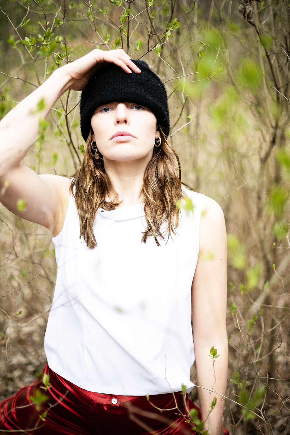 Junge Frau in weißen Oberteil und roten Hosen, trägt eine schwarze Mütze, steht in einem Busch im Freien.