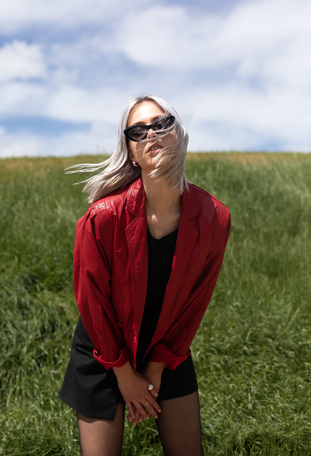 Frau mit blonden Haaren in Sonnenbrille, rotem Jacke und schwarzem Rock posiert im Grünen unter einem blauen Himmel mit Wolken.