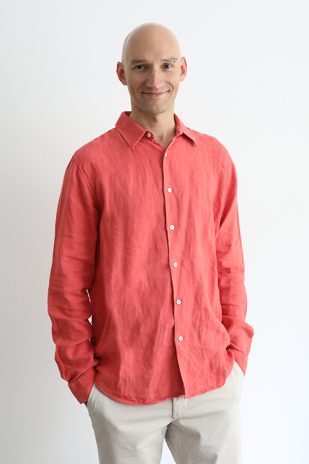 Büroangestellter trägt ein rotes Hemd und beige Hosen, lächelt, steht vor weißer Wand.