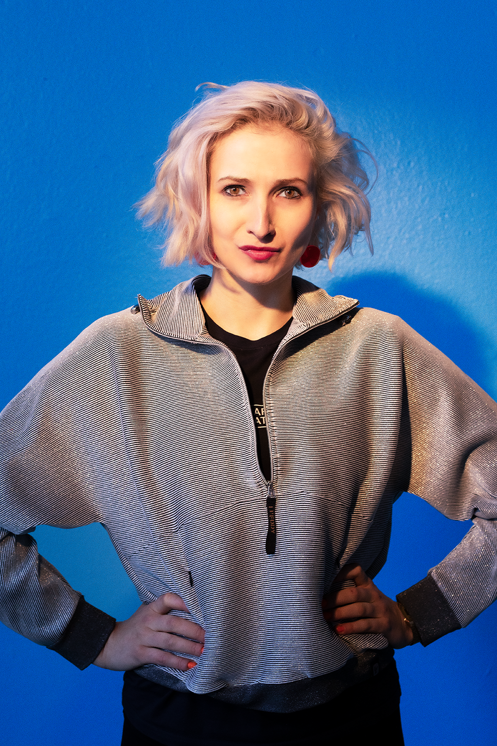 Junge Frau mit kurzen, wuscheligen, blond-pink gefärbten Haaren steht vor einem blauen Hintergrund, trägt eine silberne Jacke mit schwarzen Ärmelabschlüssen und schaut direkt in die Kamera.