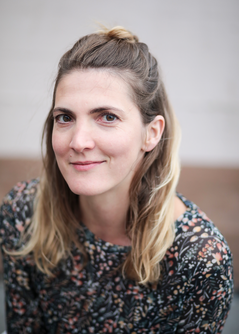 Junge Frau mit braunen, blond gefärbten Haaren, die zu einem Dutt zusammengebunden sind, sitzt lächelnd vor neutralem Hintergrund.